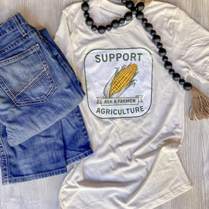 Tan tshirt that says support agriculture on it with a corn cob in the middle. Ask a Farmer